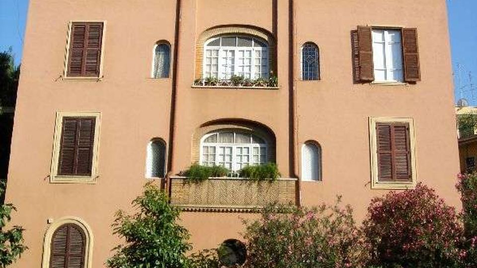 Facciata di un palazzo residenziale a più piani intonacato color ocra, con numerose finestre dotate di persiane in legno e balconi ad arco centrali. In primo piano si vedono un cancello d’ingresso con recinzione, alberi e cespugli lungo la strada.