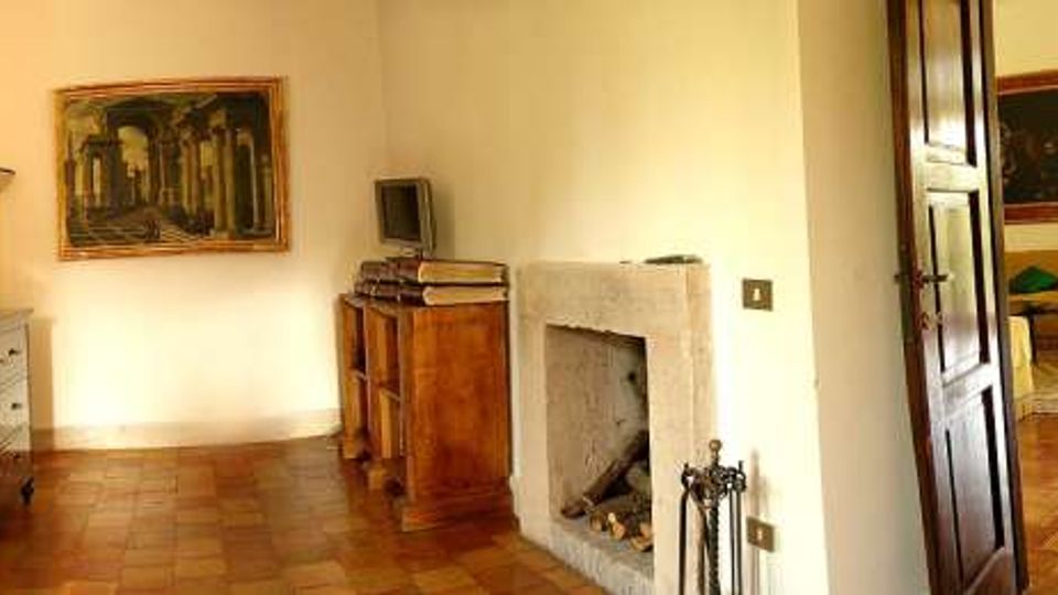 Interno di una casa con pavimento in cotto, pareti chiare e quadri incorniciati; al centro si vede un camino in pietra e mobili in legno. Una porta aperta conduce a una stanza adiacente arredata con divano e tavolo.