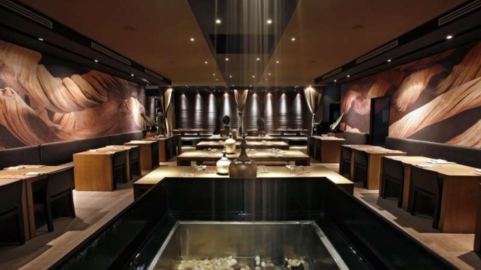 Interno di un ristorante moderno con tavoli in legno disposti in file e illuminazione soffusa a faretti sul soffitto. In primo piano c’è una vasca rettangolare con acqua e ciottoli, mentre le pareti laterali presentano grandi murales astratti e in fondo si vedono tende e pannelli scuri.