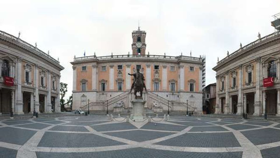 Ampia piazza lastricata con disegni geometrici, dominata da una statua equestre centrale e da un palazzo storico con facciata rosa, scalinata e torre dell’orologio. Ai lati si vedono due edifici porticati simmetrici che incorniciano lo spazio aperto.
