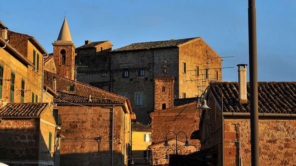 Vista di un borgo storico con case in pietra dai tetti in tegole, vicoli stretti e un campanile con guglia sullo sfondo. La luce calda del tramonto illumina le facciate, con lampioni e antenne visibili tra gli edifici.