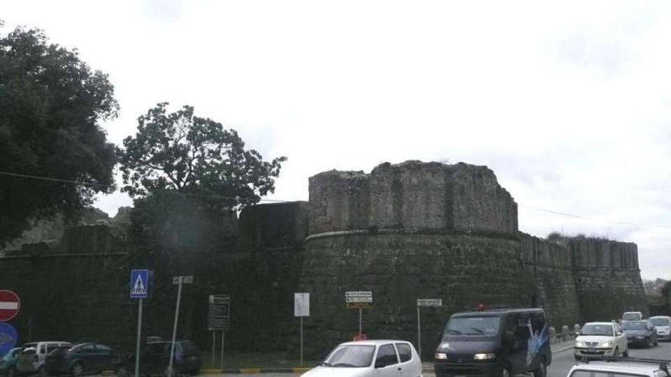 Strada urbana con traffico veicolare in primo piano, affiancata da una grande fortificazione in pietra con mura spesse e torre circolare. Sullo sfondo si vedono alberi e segnaletica stradale sotto un cielo nuvoloso.