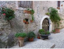 Facciata di una casa in pietra su una viuzza acciottolata, con porta ad arco in muratura e finestra con inferriate. Vasi e fioriere in terracotta con piante e fiori decorano il muro e il piccolo spazio davanti all’ingresso.