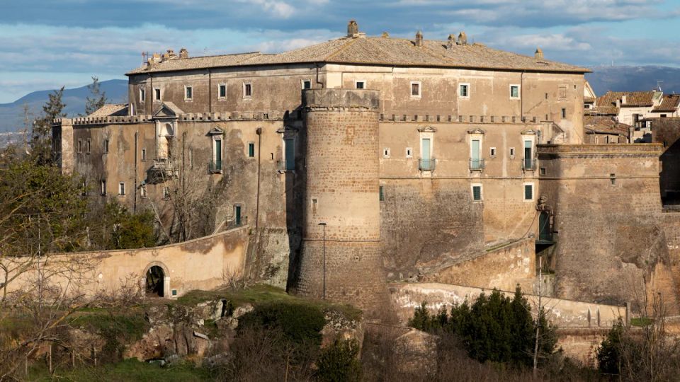 Grande edificio storico in pietra, simile a un castello o palazzo fortificato, con torre cilindrica, mura alte e finestre rettangolari. In primo piano si vede una rampa murata con arco d’ingresso, vegetazione e rovine, con colline sullo sfondo sotto un cielo nuvoloso.