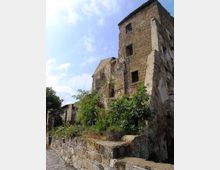Edificio in pietra in rovina con torre a più piani, finestre vuote e intonaco scrostato. In primo piano un muretto di pietra e vegetazione spontanea che cresce lungo i resti della struttura, sotto un cielo parzialmente nuvoloso.