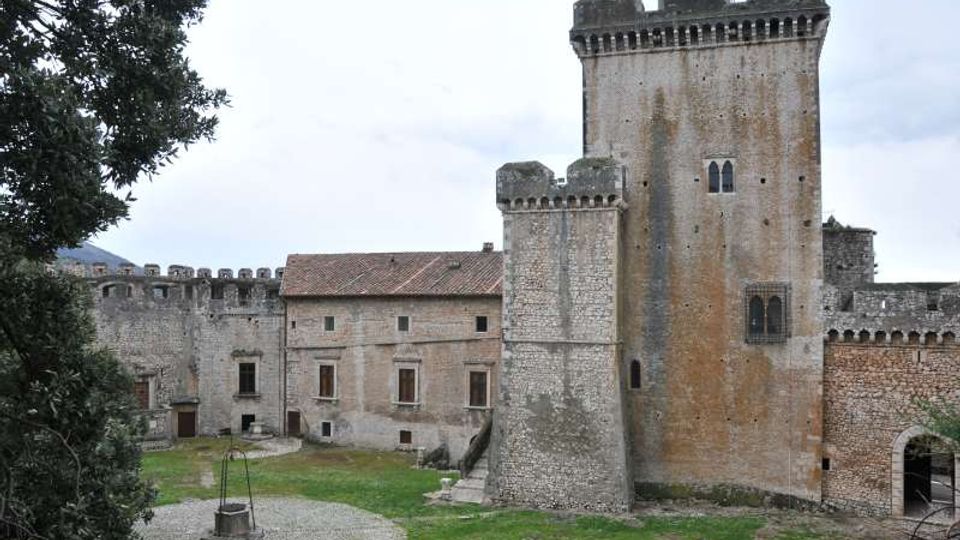 Cortile di un castello in pietra con alte torri merlate e mura fortificate, affiancate da un edificio con tetto in tegole e finestre rettangolari. In primo piano si vede un prato con un’area circolare in ghiaia e un pozzo, con un arco d’ingresso sulla destra.