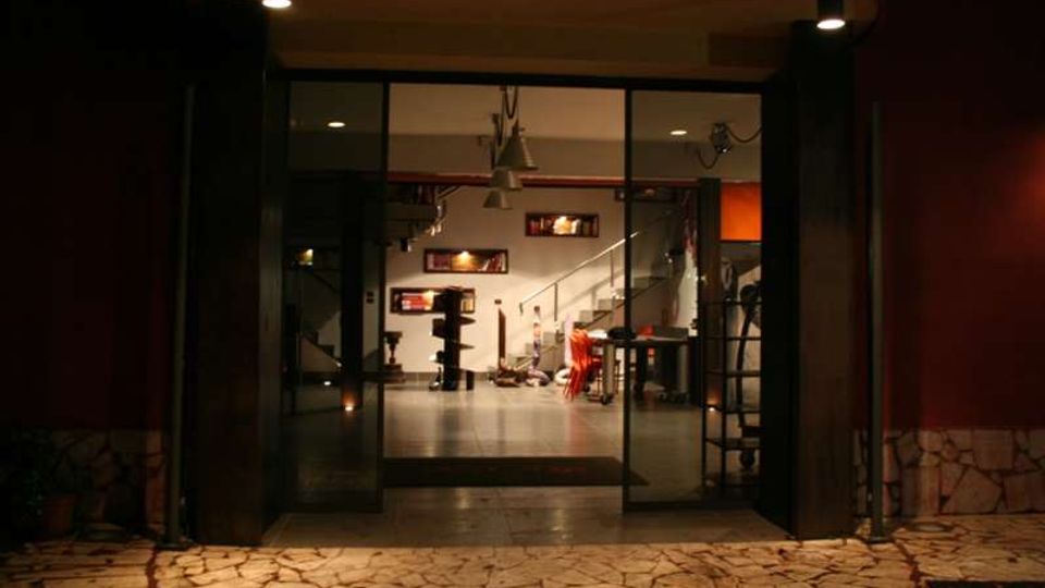 Ingresso notturno di un edificio moderno con ampia porta vetrata a tutta altezza e pavimentazione esterna in pietra irregolare. All’interno si vede un ambiente open space illuminato con scaffali a parete, arredi contemporanei e una scala con corrimano metallico che sale al piano superiore.