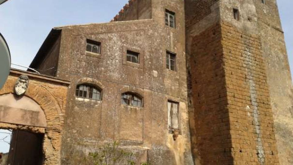 Edificio storico in muratura dall’aspetto di torre fortificata, con facciata in intonaco consumato e finestre piccole ad arco. Sulla destra si vede una torre quadrangolare in mattoni con merlature in cima, collegata a un arco d’ingresso laterale.