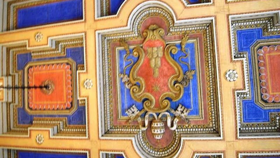 Soffitto decorato con riquadri geometrici blu e arancioni incorniciati da cornici dorate, con medaglione centrale ornato da motivi floreali e araldici. Al centro è visibile un punto luce pendente.
