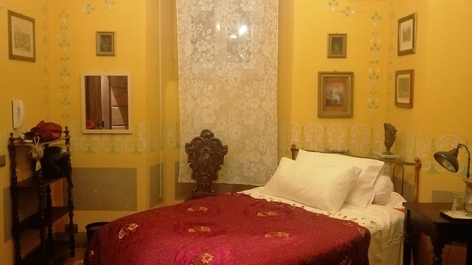 Camera da letto in stile tradizionale con soffitto a volta dipinto, pareti gialle decorate e pavimento in cotto. Un letto con coperta bordeaux è al centro sotto una finestra alta con tenda in pizzo, affiancato da comodini e quadri appesi alle pareti.