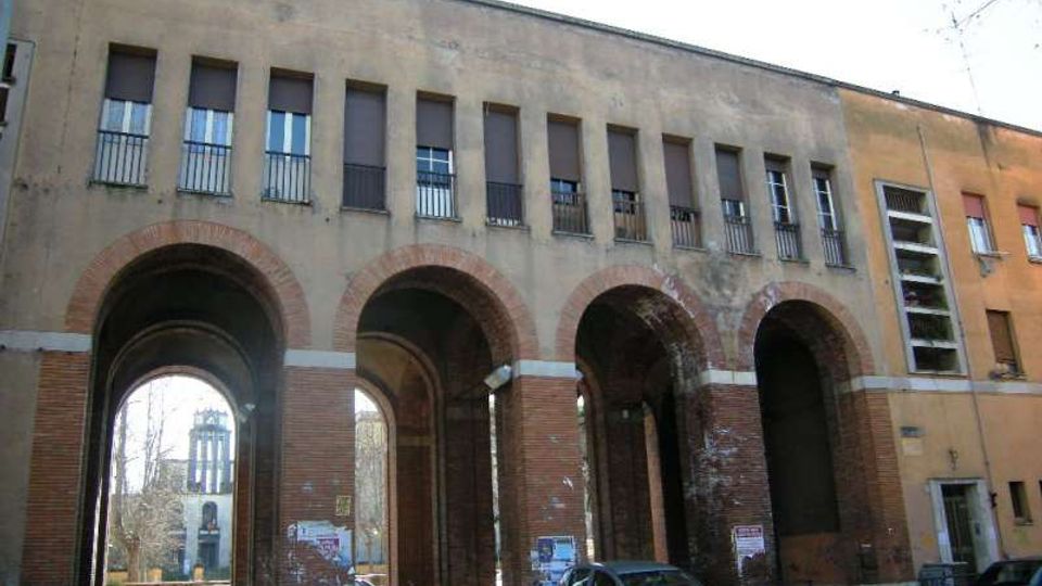 Facciata di un edificio urbano con grandi arcate in mattoni che formano un portico a più passaggi; sopra, una fila di finestre rettangolari con persiane e piccole ringhiere. Sotto le arcate si intravede la strada con auto parcheggiate e, sullo sfondo, una torre.