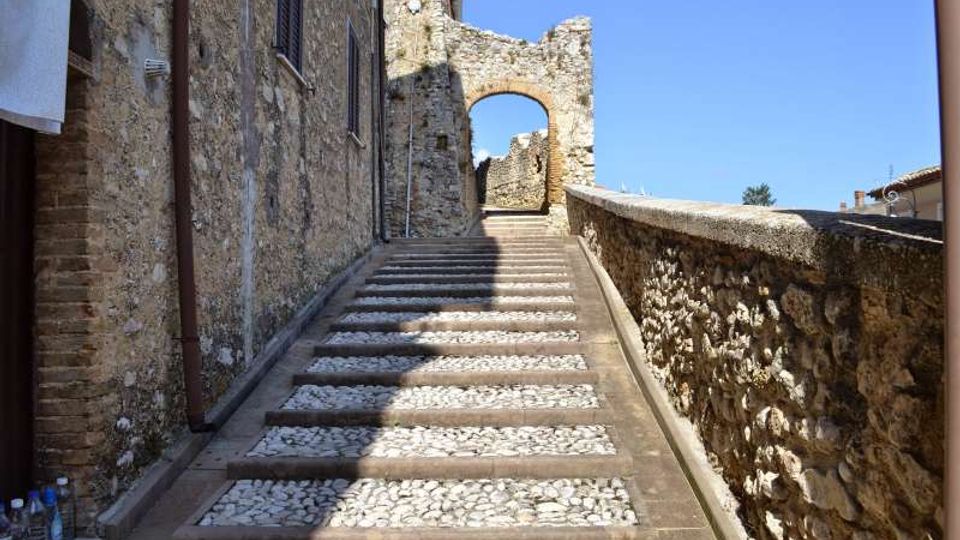 Scalinata in pietra con inserti di ciottoli bianchi che sale tra muri in pietra di edifici antichi, lungo un passaggio stretto. In cima si vede un arco in muratura collegato a una struttura di rovine, sotto un cielo limpido.