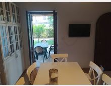 Sala da pranzo con tavolo bianco e sedie, armadi a vetri lungo la parete sinistra e TV a schermo piatto montata a destra. Una porta finestra aperta conduce a un patio con tavolo e sedie in plastica e affaccio su un giardino con prato e alberi.