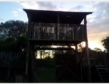 Torretta in legno rustica sopraelevata con tetto sporgente e pareti a listelli, montata su pali e accessibile tramite una scala laterale. La struttura si trova in un’area esterna recintata con alberi sullo sfondo e cielo al tramonto.