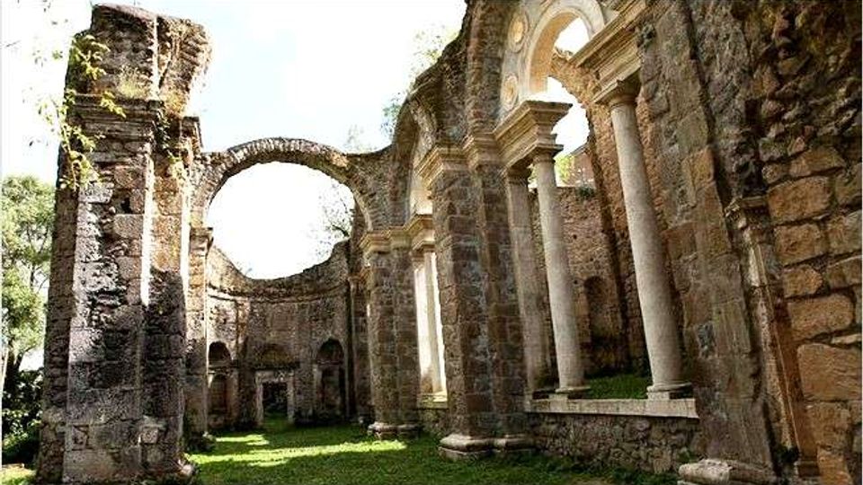 Rovine di una chiesa o abbazia in pietra, con alti muri senza tetto e grandi archi parzialmente crollati. Sul lato destro si vede un portico con colonne e un’apertura circolare sopra l’arco, mentre il pavimento interno è coperto d’erba.