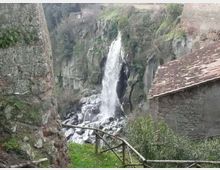 Cascata naturale che scende da una parete rocciosa in una gola, con massi alla base e vegetazione sulle scarpate. In primo piano si vedono un sentiero erboso con staccionata in legno e muri in pietra, con un edificio in pietra dal tetto in tegole sulla destra.