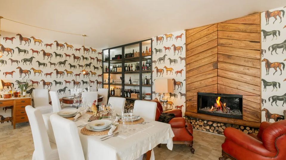 Sala da pranzo rustica con grande tavolo apparecchiato e sedie rivestite di bianco, pareti con carta da parati a cavalli e vetrina con bottiglie. A destra c’è un camino acceso incorniciato da pannelli di legno a spina di pesce, con legnaia sottostante e poltrone in pelle rossa.