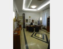 Interno di un ufficio direzionale con grande scrivania in legno, sedie nere e pareti decorate con cornici, attestati, stemmi e una bandiera vicino alla finestra con tende bianche. Pavimento in marmo chiaro con intarsi geometrici scuri e soffitto ribassato con illuminazione integrata.