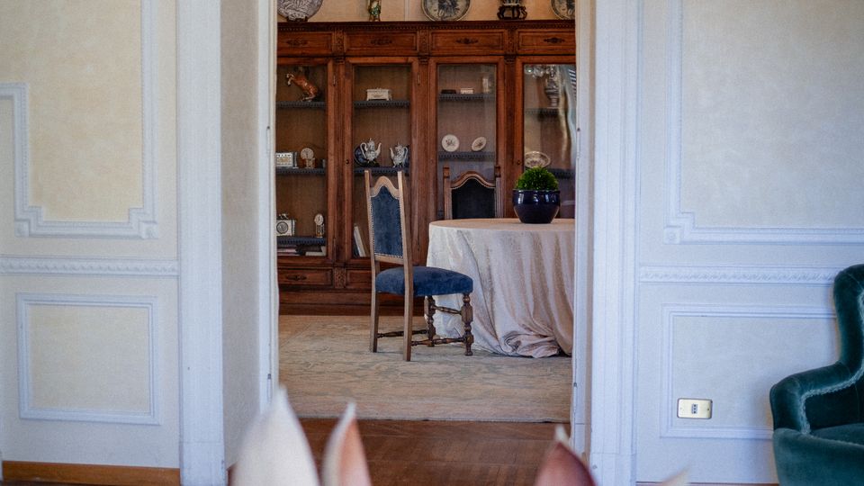 Vista attraverso una porta interna di un salone classico con pareti chiare modanate e pavimento in parquet, che conduce a una sala da pranzo. Nella stanza sullo sfondo si vedono un lampadario in cristallo, una grande credenza in legno con stoviglie esposte e un tavolo rotondo coperto da una tovaglia.