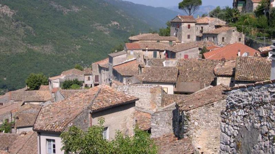 Vista di un borgo collinare con case in pietra addossate tra loro e tetti in coppi di terracotta. Sullo sfondo si vedono pendii montuosi verdi e un albero isolato sopra le abitazioni.