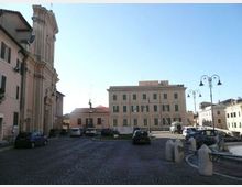 Piazza lastricata in pietra con auto parcheggiate, circondata da edifici storici: a sinistra una chiesa con facciata alta e decorata, di fronte un palazzo rettangolare con persiane verdi. Lampioni in ferro e una piccola area con balaustra in primo piano definiscono il bordo della piazza sotto un cielo sereno.