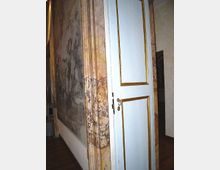 Interno di un edificio storico con pareti affrescate e rivestimenti in marmo venato; al centro una porta bianca con cornici dorate e maniglia in ottone. Pavimento in legno scuro e corridoio che prosegue sullo sfondo.