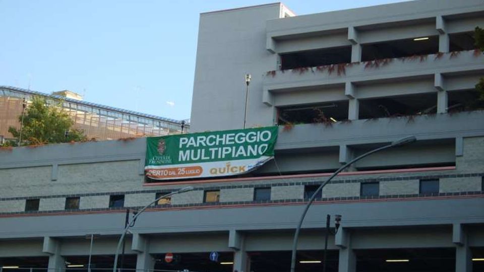Parcheggio multipiano in cemento grigio con rampe e aperture orizzontali su più livelli, visto dall’esterno in ambiente urbano. Sulla facciata è appeso un grande striscione verde con la scritta “PARCHEGGIO MULTIPIANO”, con lampioni e strada in primo piano.