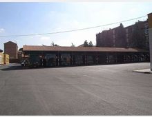 Ampio piazzale asfaltato con una lunga tettoia coperta che ospita una fila di autobus parcheggiati. Sullo sfondo si vedono edifici residenziali in mattoni e alcuni alberi, con cavi elettrici sospesi sopra l’area.