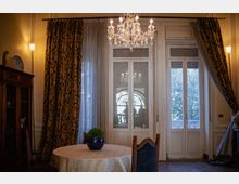 Interno di una sala da pranzo in stile classico con lampadario in cristallo, tavolo rotondo e mobili in legno. Grandi porte-finestre bianche con tende pesanti e leggere si affacciano su un balcone con alberi visibili all’esterno.