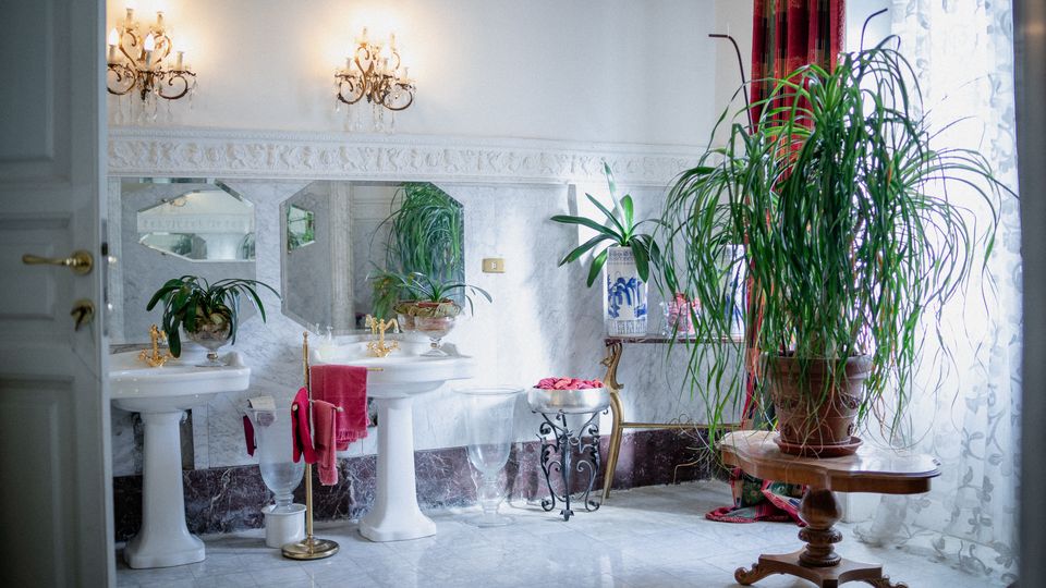 Bagno ampio e luminoso con pavimento in marmo, due lavabi su colonna con rubinetteria dorata e specchi a parete, illuminati da applique classiche. Vicino a una grande finestra con tende e drappeggi rossi, un tavolino in legno sostiene una grande pianta in vaso.