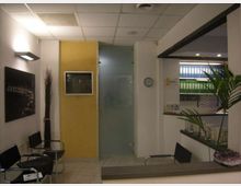 Interno di una sala d’attesa in ufficio, con sedie nere lungo la parete, pavimento piastrellato e controsoffitto a pannelli con luci. Si vedono una parete gialla con display incassato, una porta in vetro satinato, un bancone con scaffali di raccoglitori e alcune piante decorative.