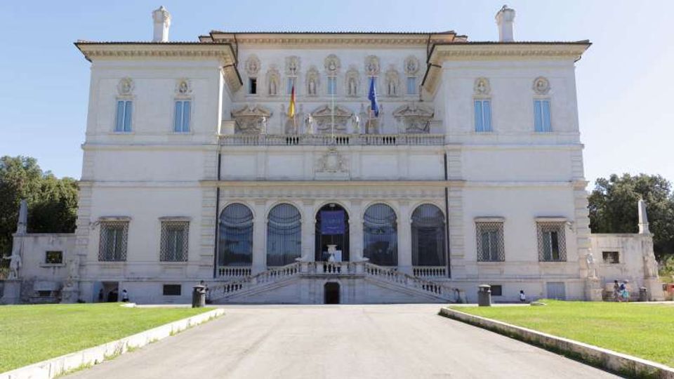 Facciata di un grande palazzo neoclassico chiaro, simmetrico, con finestre alte e tre arcate centrali al piano terra, raggiungibili da una scalinata con balaustra. Davanti si estende un viale rettilineo tra prati verdi, con bandiere sul balcone centrale e alberi ai lati sullo sfondo.