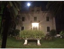 Edificio storico a due piani illuminato di notte, con ingresso centrale ad arco e finestre simmetriche sulla facciata. Davanti si vede un giardino con prato, aiuola rialzata e palme ai lati, sotto un lampione.