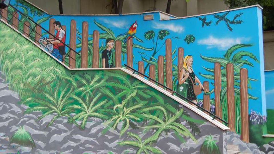 Scala esterna in cemento con corrimano metallico, affiancata da un grande murale dai colori vivaci che raffigura vegetazione tropicale, pali di legno e un pappagallo su sfondo azzurro. Il murale prosegue lungo il muro laterale e segue l’inclinazione delle rampe della scala.