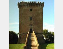 Torre in mattoni a pianta quadrata con sommità merlata e piccole finestre, raggiunta da una lunga scalinata centrale che conduce all’ingresso sopraelevato. La struttura è circondata da un prato verde con alberi ai lati sotto un cielo sereno.