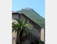 Edificio moderno in mattoni con finestre rettangolari e una palma in primo piano, ai piedi di una collina verde. In cima alla collina si vede un grande complesso monumentale chiaro, simile a un santuario o monastero, contro un cielo azzurro.