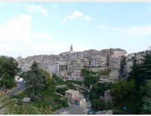 Vista panoramica di un borgo collinare con case in pietra addossate tra loro e un campanile che svetta al centro. In primo piano si vedono vegetazione, piccoli edifici rurali e una strada curvilinea ai margini del paese.