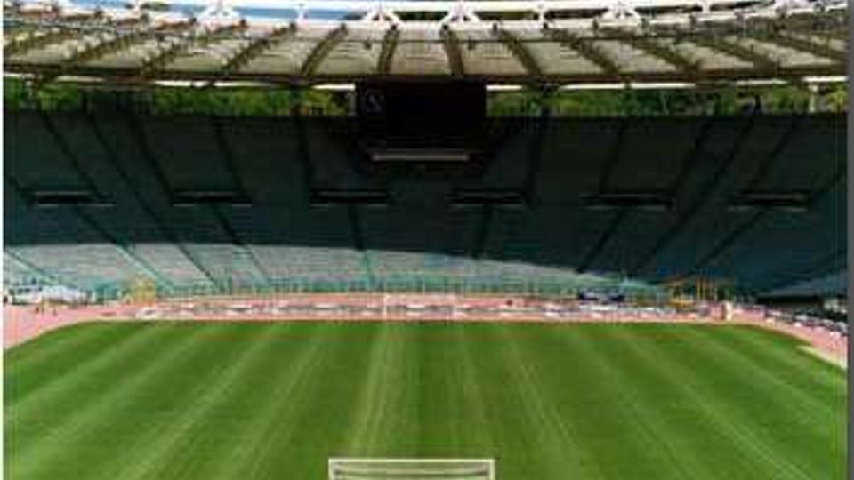 Interno di uno stadio all’aperto con ampie tribune a gradoni e copertura superiore, visto dal lato del campo. In primo piano si vede il prato verde con le linee di gioco e una porta da calcio, circondato da una pista di atletica.