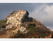 Borgo collinare con case in pietra e intonaco addossate tra loro, arroccato sulla sommità di un rilievo boscoso. Sullo sfondo un cielo nuvoloso, con vegetazione autunnale che ricopre i versanti.