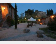 Cortile di un casale in pietra al crepuscolo con vialetti di ghiaia, luci esterne calde e un gazebo bianco al centro. Sullo sfondo si vedono colline alberate, cipressi e un edificio in pietra su un’altura.