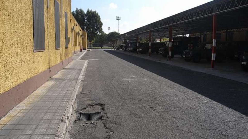 Cortile asfaltato tra un lungo edificio giallo con finestre strette e un’ampia tettoia metallica con pilastri, simile a un deposito o autorimessa. Sulla strada si notano crepe e una griglia di scolo, con alberi e lampioni sullo sfondo.