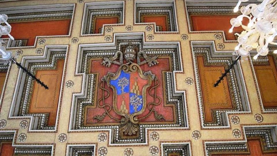 Soffitto decorato di un edificio storico con motivi geometrici a riquadri in tonalità rosso e oro, cornici ornate e dettagli a rosette. Al centro è presente uno stemma araldico in rilievo, con lampadari bianchi ai lati.