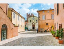 Piazza acciottolata in un borgo storico, circondata da edifici dai colori pastello con persiane e facciate in intonaco. Sullo sfondo si vede una piccola chiesa con portale centrale, orologio sulla facciata e campanile in pietra.