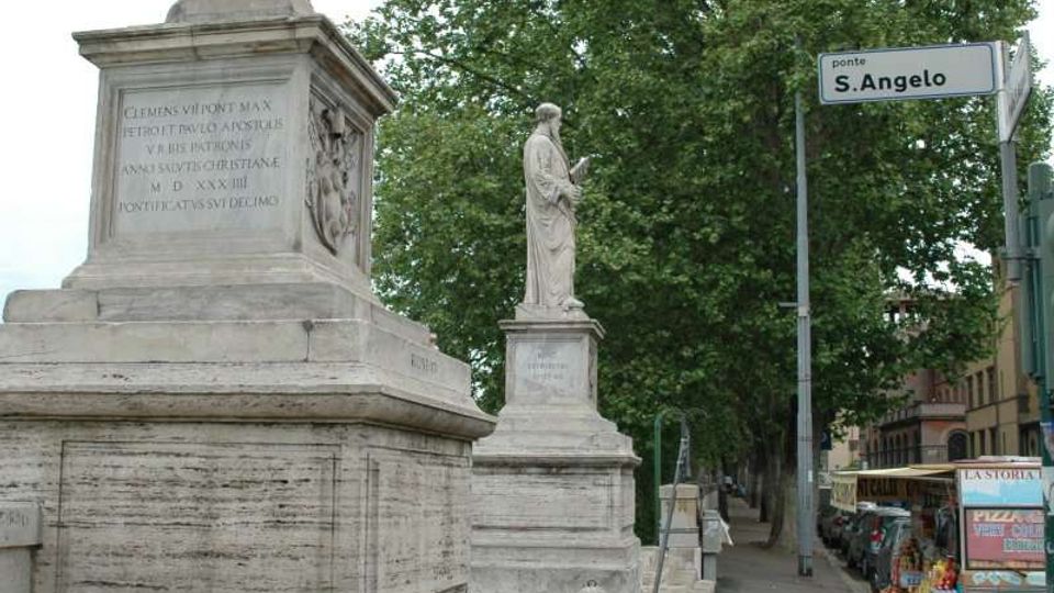 Statue in pietra su alto basamento con gradini all’ingresso di Ponte Sant’Angelo, con un’altra statua allineata lungo il parapetto. A destra si vede il cartello stradale “ponte S. Angelo”, una fila di alberi e una strada con bancarelle.