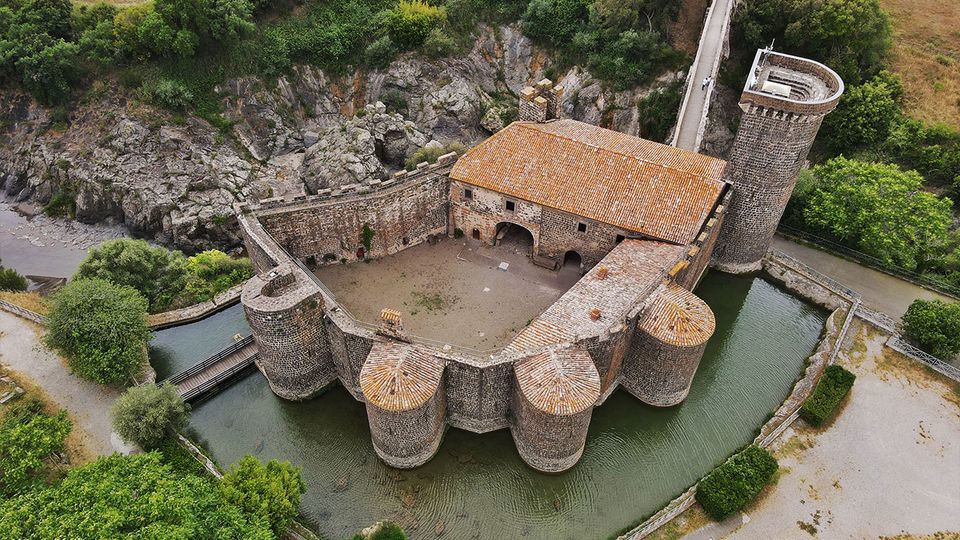 Veduta aerea di un castello in pietra circondato da un fossato d’acqua, con mura e torri cilindriche e un edificio centrale con tetto in tegole. Intorno si vedono un ponte d’accesso, una strada e aree verdi e campi coltivati ai margini.