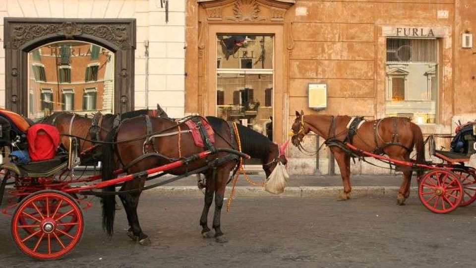 Strada urbana con pavimentazione in pietra davanti a una facciata di edifici storici con grandi portali e finestre. In primo piano ci sono carrozze trainate da cavalli con ruote rosse parcheggiate lungo il bordo della strada.