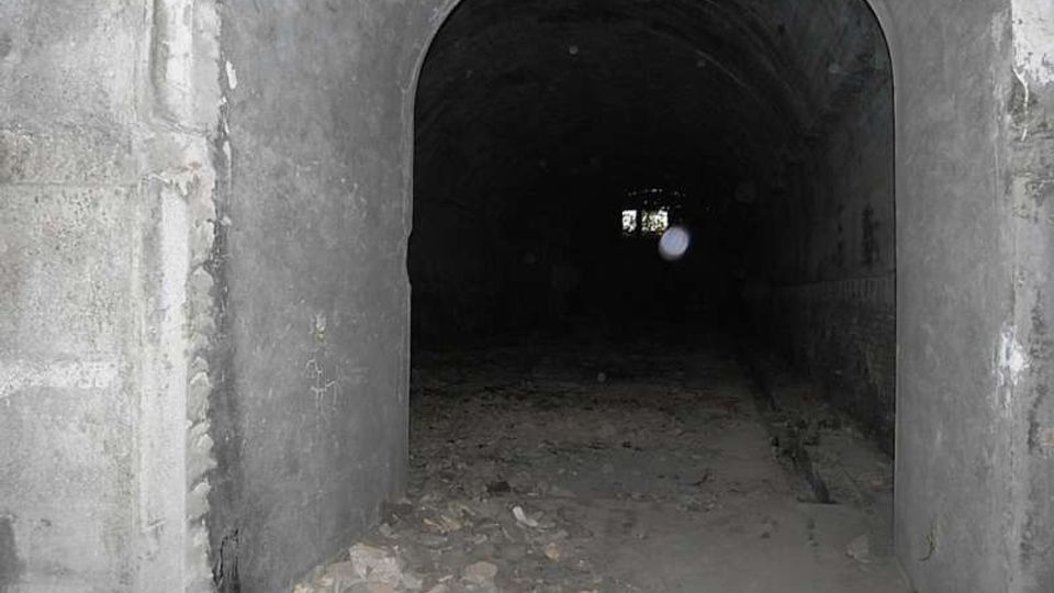 Ingresso ad arco di un tunnel sotterraneo in cemento, con pareti grezze e pavimento coperto di detriti. Il corridoio è buio e si intravede una piccola apertura luminosa in fondo.