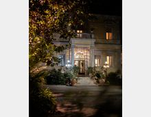 Facciata notturna di una villa storica con ingresso ad arco, cancellata in ferro battuto e scalinate illuminate da lampioni. Davanti si estende un giardino con piante ornamentali e vasi ai lati dei gradini.