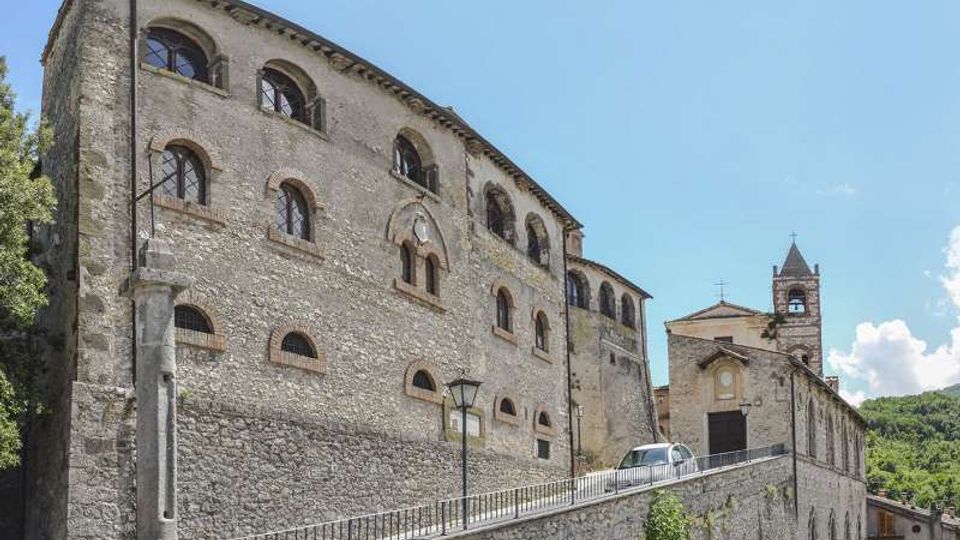 Complesso storico in pietra su un pendio, con grande edificio a più piani e finestre ad arco, collegato da una rampa con parapetto metallico. A destra si vede una chiesa in pietra con campanile e facciata semplice, sullo sfondo colline verdi e cielo sereno.