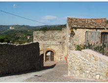 Stradina in pietra in discesa tra muri in pietra e case rustiche con tetto in coppi, con un portale ad arco in mattoni e una lanterna a parete. Sullo sfondo si vedono colline verdi e campagna sotto un cielo azzurro.
